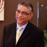 A smiling man in a black suit and pale yellow tie stands against a neutral background, with abstract art and greenery visible nearby. His glasses and confident expression convey professionalism and warmth.