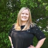 A smiling young woman stands outdoors in front of a leafy green background. She wears a black shirt adorned with decorative beads and light-colored pants, posing with her hands on her hips.