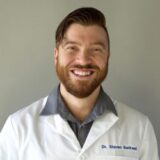A smiling medical professional with a beard, wearing a white lab coat over a dark shirt, poses against a plain background.