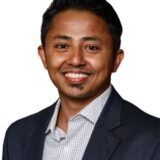 A smiling man with short black hair, wearing a dark blazer over a light-patterned shirt. He has a goatee and is positioned against a plain white background.