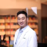 A smiling man in a white lab coat stands in a clinical setting with colorful shelves in the background, suggesting a professional healthcare environment.