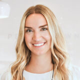 A smiling woman with long, blonde hair is standing in a well-lit environment, wearing a light top. She exudes warmth and friendliness, showcasing a welcoming spirit perfect for her role in a dental setting.