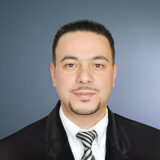 A professional-looking man with short dark hair and a light beard, wearing a black suit and a striped tie, smiles confidently against a neutral backdrop.
