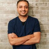 A smiling man stands with his arms crossed in front of a gray brick wall, wearing a navy blue shirt. The setting conveys a casual and friendly atmosphere.