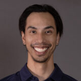 A smiling individual with dark hair and earrings is wearing a navy blue shirt. Their cheerful expression conveys friendliness and approachability, representing a welcoming atmosphere.