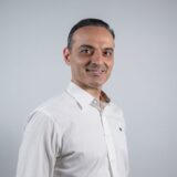 A man with short dark hair and a friendly smile is wearing a white collared shirt. He is facing slightly to the right against a light gray background.