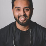 A smiling dental professional wearing a black coat and dental tools around his neck. The background is neutral, emphasizing his friendly demeanor.