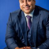 A man dressed in a navy suit and purple tie smiles confidently while seated against a blue background.