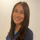 A smiling woman with long, dark hair poses in a casual blue outfit, standing against a neutral background.