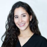 A young woman with long, curly hair smiles warmly, wearing a black top. The background is a soft, light color, enhancing her friendly demeanor.
