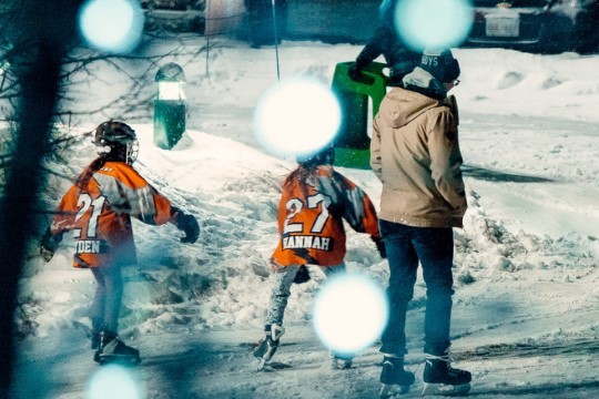 Two children in orange jerseys skate on a snowy rink, while an adult stands nearby. The scene is slightly blurred with glowing blue orbs in the foreground, suggesting a cold, winter evening atmosphere.
