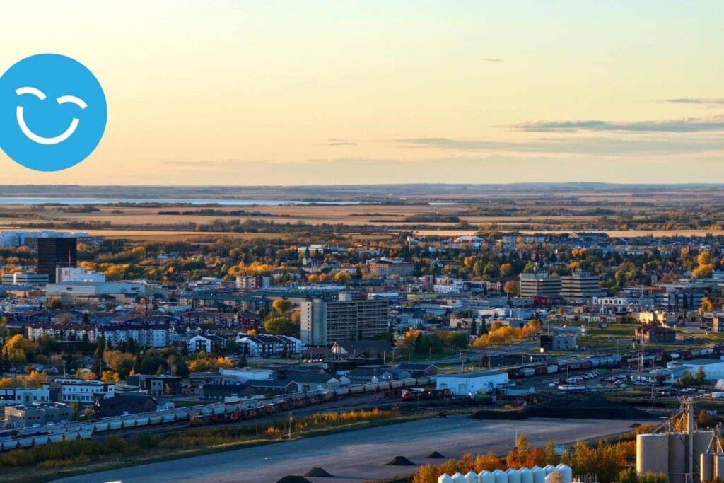 A scenic aerial view of a city at sunset, showcasing buildings and green spaces, with a river in the foreground and a soft, warm light illuminating the landscape. A blue smiley face icon appears in the corner.