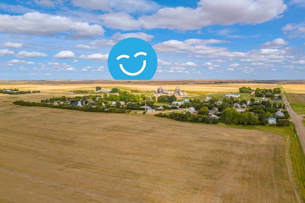 Aerial view of a small rural community surrounded by expansive fields and blue sky. A large blue smiley face icon appears in the sky above the town, suggesting a cheerful atmosphere.