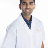 A smiling man wearing a white lab coat over a dark polo shirt stands against a plain white background, suggesting a professional or medical context.