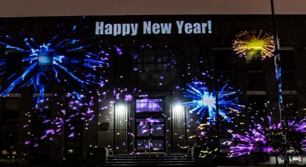A building is illuminated with colorful projections of fireworks, with the words "Happy New Year!" displayed prominently at the top. The scene is set at night, adding a festive atmosphere to the celebration.