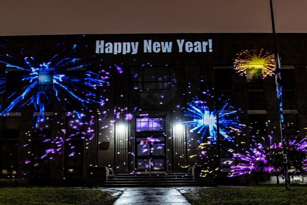 A building is illuminated with colorful projections of fireworks, with the words "Happy New Year!" displayed prominently at the top. The scene is set at night, adding a festive atmosphere to the celebration.