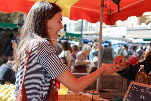 A smiling woman engages with a vendor at a bustling market under bright red umbrellas. She is exchanging money for goods, surrounded by colorful stalls and shoppers enjoying the lively atmosphere.
