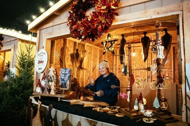 A vendor stands behind a wooden stall adorned with handcrafted ornaments and decorations, including wooden figurines and a wreath. The setting is festive, with warm lighting highlighting the charming display.