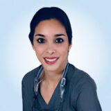 A smiling dental professional in gray attire poses against a light blue background, showcasing her friendly demeanor and approachability in a clinical setting.