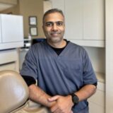 A dental professional in scrubs stands with a confident smile, leaning against a dental chair in a modern office setting. The background features cabinets and equipment, highlighting a clinical environment.