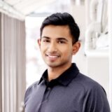 A young man with short black hair smiles while standing indoors, wearing a dark polo shirt. He appears confident and friendly, with soft natural light highlighting his features.