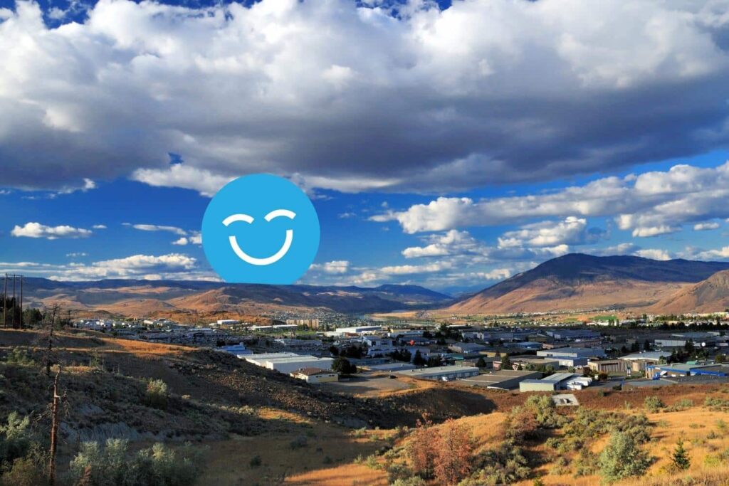 A panoramic view of a valley bordered by mountains under a partly cloudy sky. In the foreground, a mix of grassy and rocky terrain leads to a small industrial area. A blue smiley face icon overlays the image.