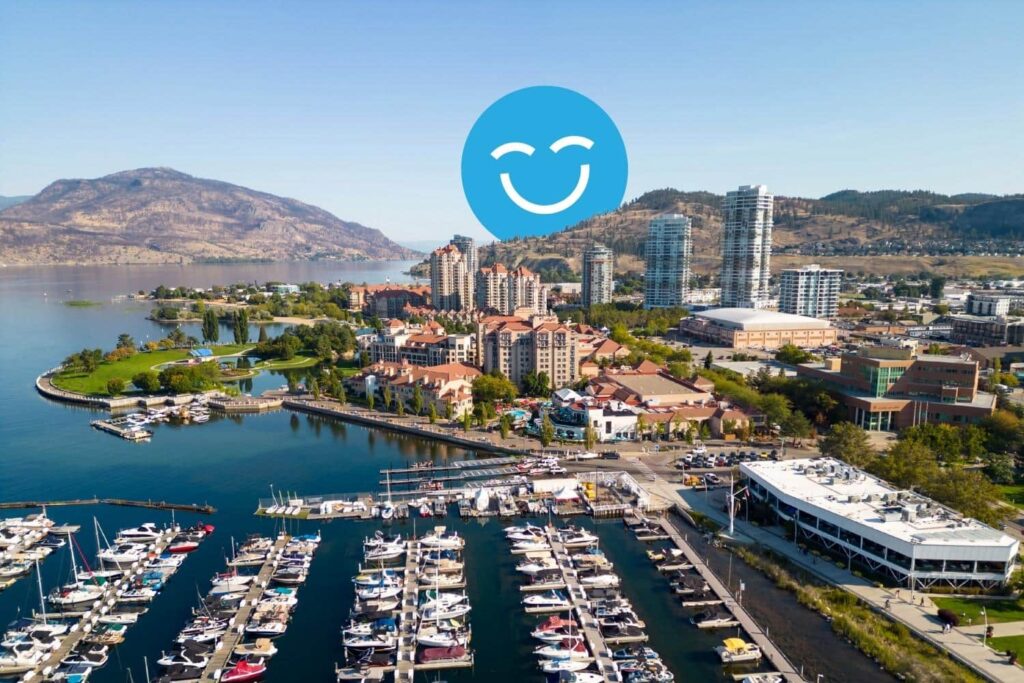 Aerial view of a vibrant waterfront marina surrounded by modern buildings, with boats docked in the harbor and mountains in the background. A cheerful blue smiley face icon overlays the scene, adding a playful touch.