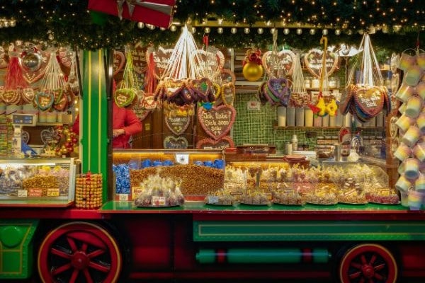 A festive market stall is filled with colorful treats and candies, including heart-shaped confections. The stall is adorned with greenery and lights, creating a cheerful holiday atmosphere. In the background, a vendor is seen preparing items, enhancing the lively scene of holiday shopping.