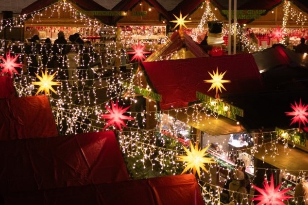 A vibrant market scene illuminated by colorful lights and stars, featuring festive decorations on wooden stalls and rooftops. The atmosphere is lively, suggesting a celebration or holiday event.