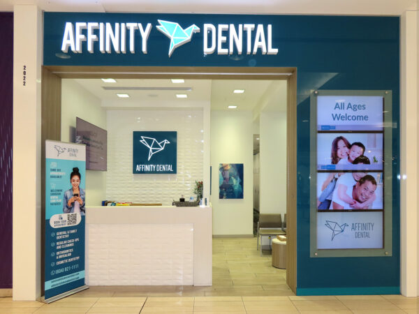 Entrance of Affinity Dental clinic featuring a reception desk, blue and white decor, and promotional screens displaying family-friendly dental services. A banner invites all ages to visit.