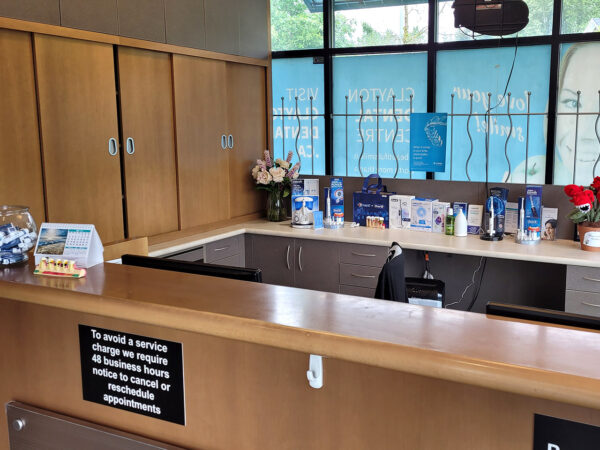 Un espace d'accueil avec un comptoir en bois, un bol en verre de bonbons, diverses brochures et des fleurs décoratives. De grandes fenêtres laissent entrer la lumière naturelle, et une enseigne est visible sur le comptoir.