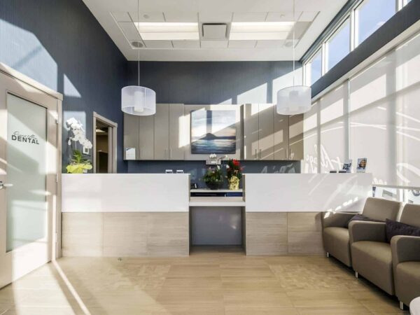 A modern reception area featuring a sleek white desk, comfortable seating, and large windows allowing natural light. Decorative plants enhance the welcoming atmosphere. The front door is labeled "Dental."