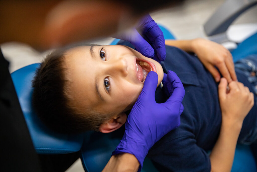 A young boy sits in a dental chair, smiling as a dental professional examines his teeth. The dentist wears purple gloves and gently holds the boy's chin while checking his mouth. A friendly blue smiley face, the 123Dentist Smiley, adds a cheerful touch to the scene.