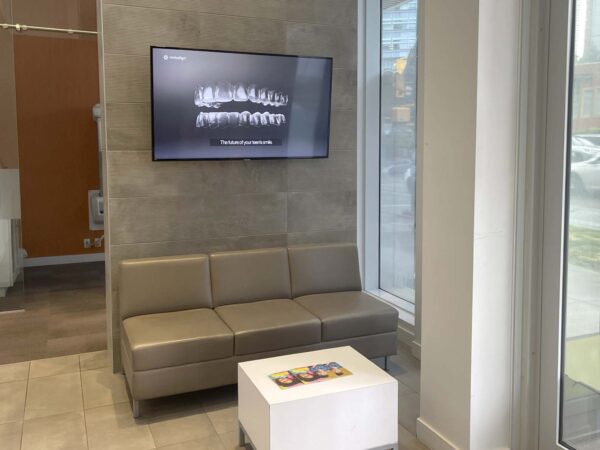 A waiting area featuring a light gray couch, a small white table displaying magazines, and a wall-mounted screen showing dental images. Large windows allow natural light into the space.