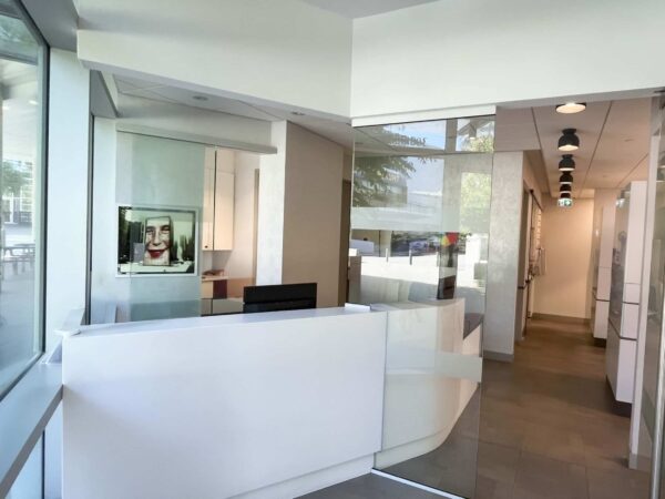 A modern office reception area features a white reception desk, glass walls, and a hallway leading to other office spaces. Natural light illuminates the area, and a large portrait is visible on the wall.