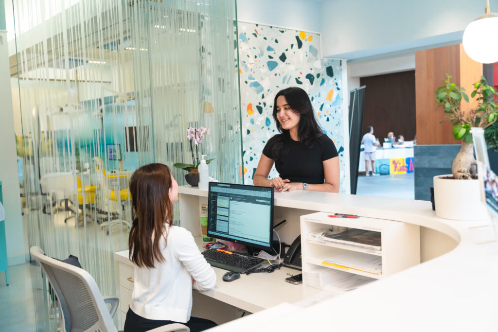 A reception area where a woman in a black shirt engages with a seated staff member at a desk with a computer. The space features glass partitions and a bright, modern design.