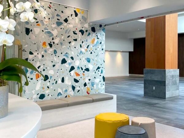 A modern interior space featuring a colorful terrazzo wall, a light-colored bench, and decorative plants. The area includes round seating in yellow and gray tones, with wooden and stone accents visible in the background.
