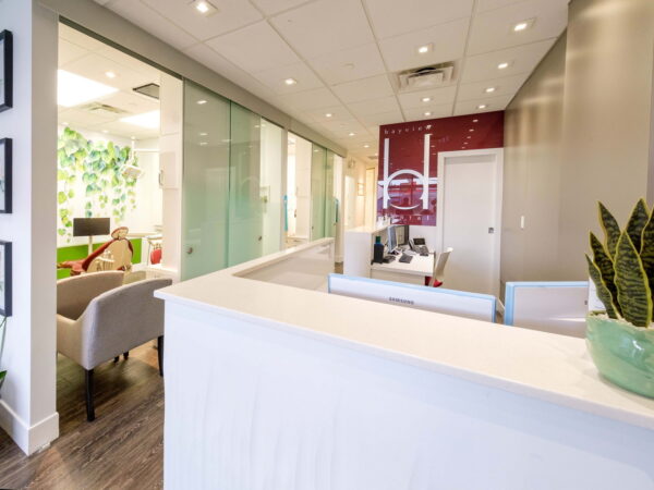 A modern reception area featuring a sleek white desk, contemporary decor, and glass partitions. Green plants are placed throughout, adding a touch of nature to the inviting space. The background shows comfortable seating and an office environment.