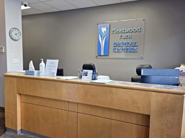 A modern dental office reception area featuring a wooden front desk, a wall clock, and informational signage. The space is clean and welcoming, designed for patient check-in and inquiries.