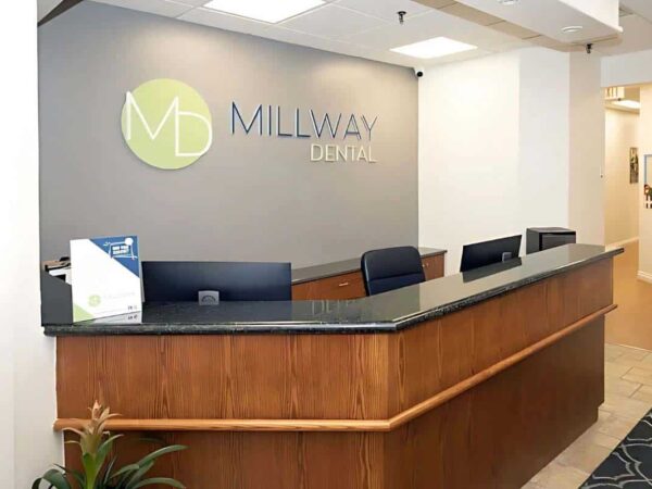 A modern dental office reception area featuring a wooden front desk, multiple black chairs, and a sign reading "Millway Dental" on a gray wall. The space is well-lit and clean, creating a welcoming environment for patients.