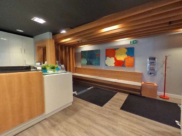 A modern and welcoming reception area featuring a low wooden countertop, colorful abstract artwork on the walls, and a seating bench. The floor is wooden with two black mats near the entrance, creating a spacious and inviting atmosphere.