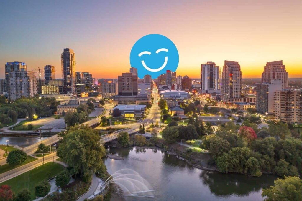 A scenic view of a city skyline at sunset, with tall buildings reflecting the warm colors of the sky. A river and green trees are in the foreground, and a smiling blue icon overlays the image.