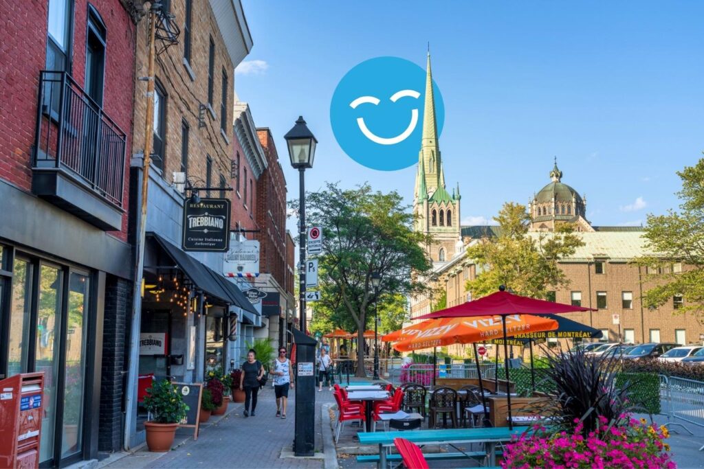 A vibrant street scene features outdoor seating with colorful umbrellas, lined with shops and greenery. In the background, a tall church steeple rises against a clear blue sky, adding to the lively atmosphere.