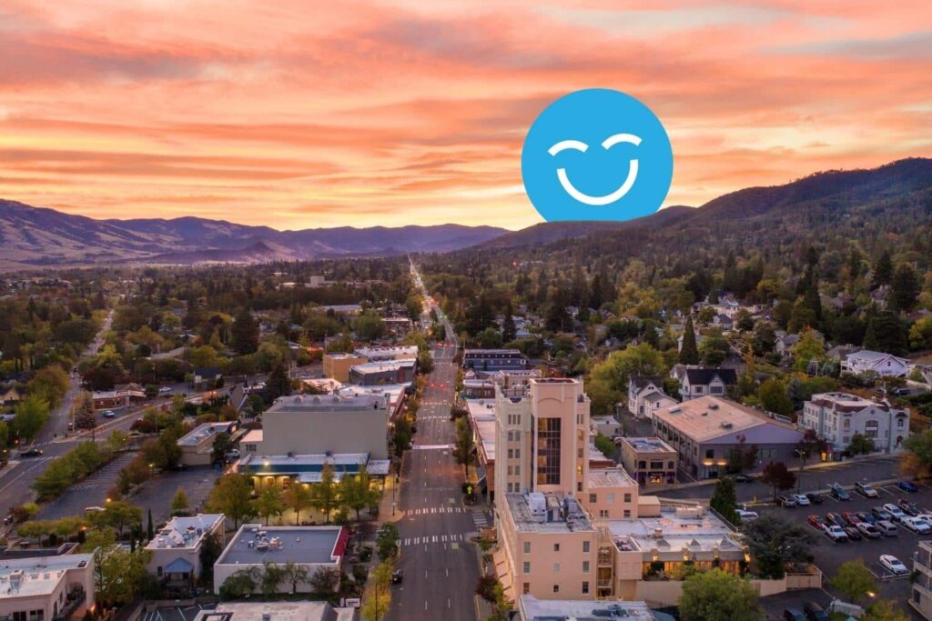 A scenic aerial view of a suburban town at sunset, with vibrant colors in the sky. A large, blue smiley face graphic is superimposed over the landscape, adding a whimsical touch to the serene setting.