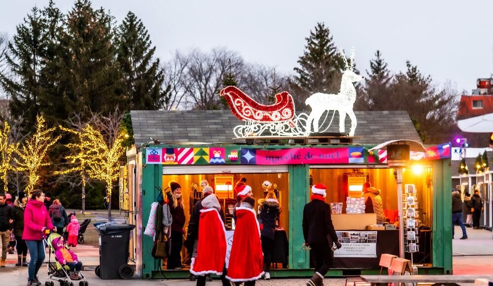 A festive market stall decorated with colorful lights, featuring a sleigh and a horse silhouette. People in Santa costumes gather outside, and families stroll nearby, enjoying the holiday atmosphere among decorated trees.