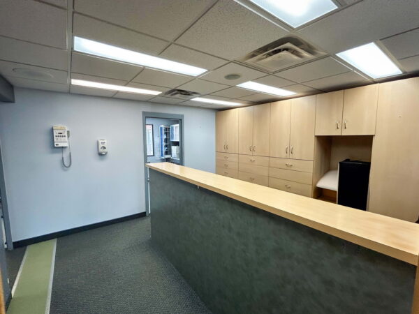 A clean, modern office reception area featuring a wooden front desk, light-colored cabinets, and a telephone on the wall. The space is well-lit with overhead fluorescent lights and has a neutral color scheme.