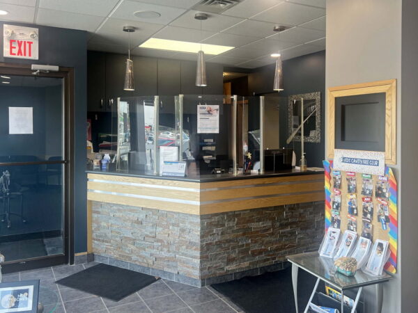 A modern reception area featuring a wooden front desk with stone accents, surrounded by pendant lights. There are informational brochures on a table nearby and a glass door leading outside. The color scheme is neutral with dark walls and a bright, welcoming atmosphere.