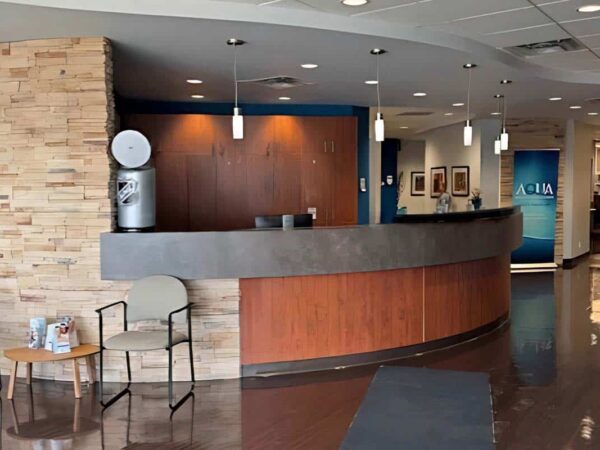 A modern reception area featuring a curved wooden front desk, stone accents, and pendant lighting. Two chairs are placed beside a small table, and a blue wall with framed art is visible in the background.