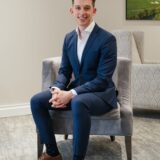 A professional young man sits comfortably in a stylish chair, wearing a navy suit, white shirt, and brown dress shoes, with a relaxed expression. A green landscape painting is visible in the background.