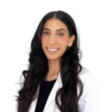 A smiling woman with long, dark hair wears a white lab coat over a black shirt. She appears friendly and approachable, set against a plain white background.